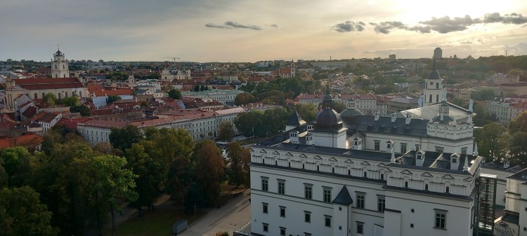A panoramic view of a cityscape featuring a mix of historical and modern architecture, with red-tiled roofs, green trees, and a cloudy sky at sunset.