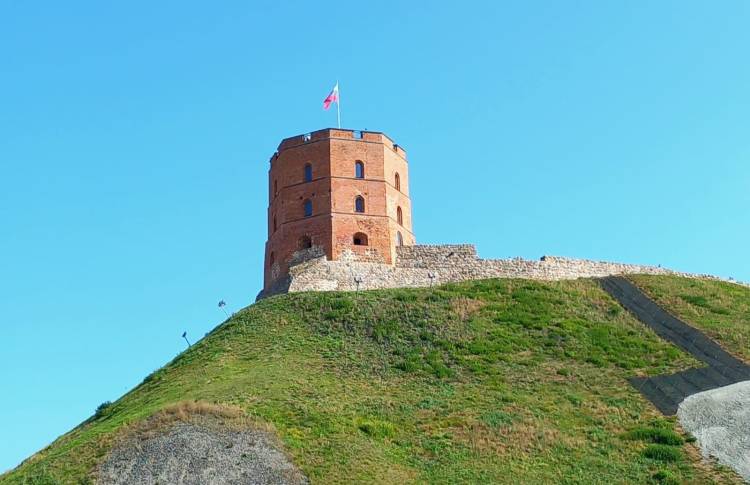 View of a historic brick tower on a grassy hill against a clear blue sky, with a flag flying at the top.