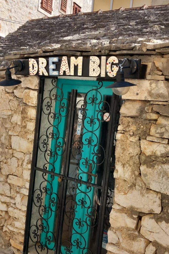 A rustic stone building entrance with a black wrought iron gate and turquoise door, featuring a sign that reads 'DREAM BIG' in colorful letters.