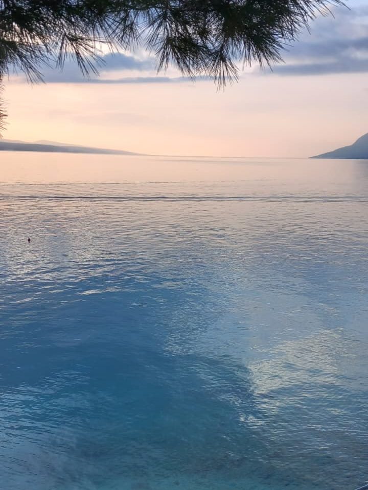 A serene view of calm water at sunset, with soft pastel colors in the sky and distant mountains in the background.