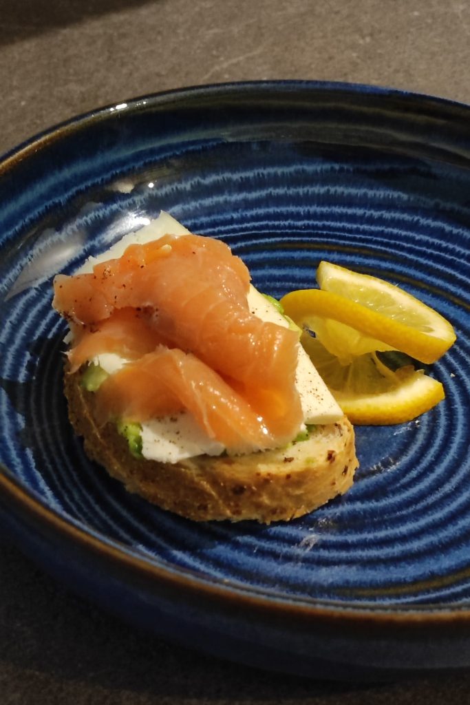 A slice of bread topped with cream cheese, avocado, and smoked salmon, served on a blue patterned plate with lemon wedges on the side.