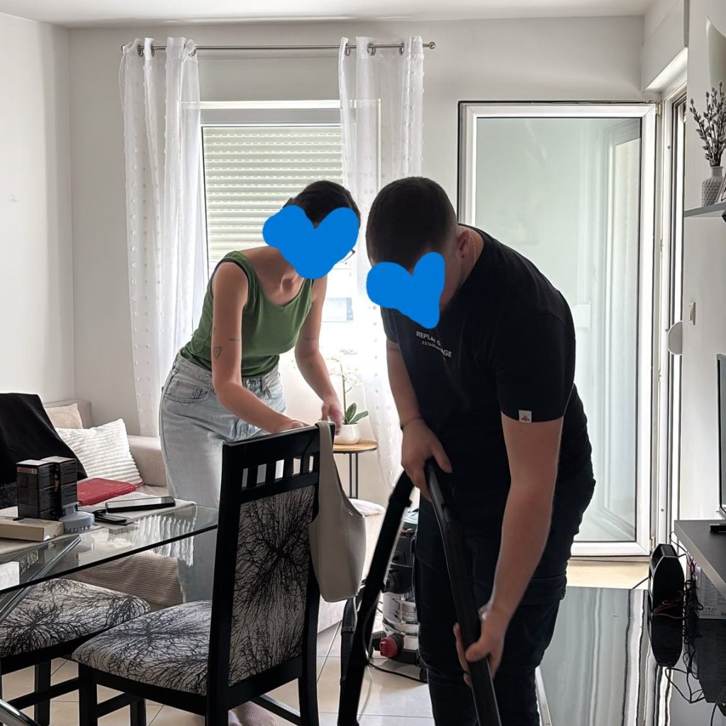 Two individuals cleaning a living room, one vacuums while the other organizes items on a table. Bright interior with large windows.
