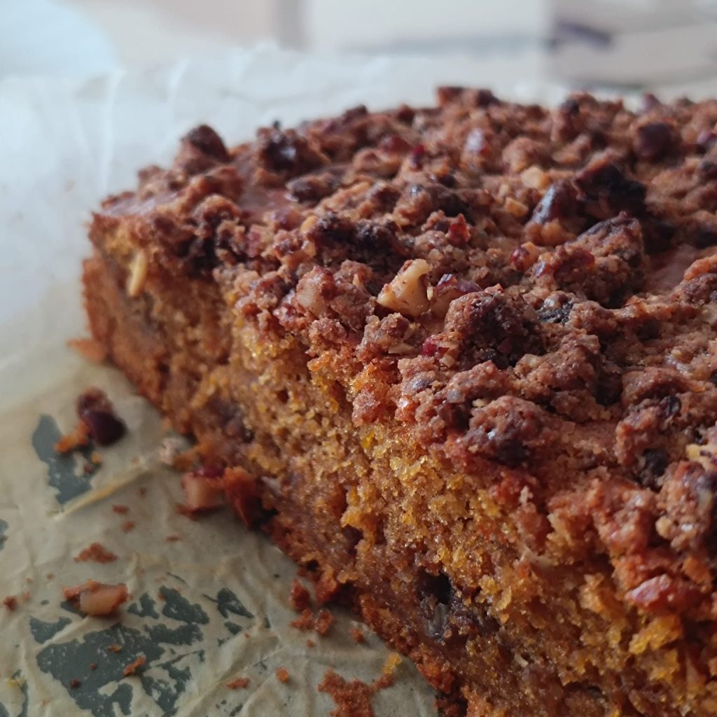 Close-up view of a slice of cake with a crumbly topping, featuring bits of nuts and a golden-brown crust on a parchment paper.