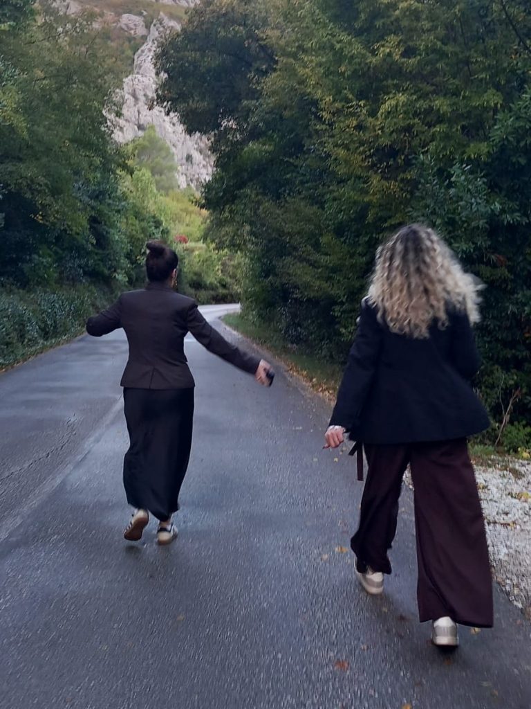 Two women walking down a tree-lined road, one is in a black suit and the other in casual clothing, both with their backs to the camera.