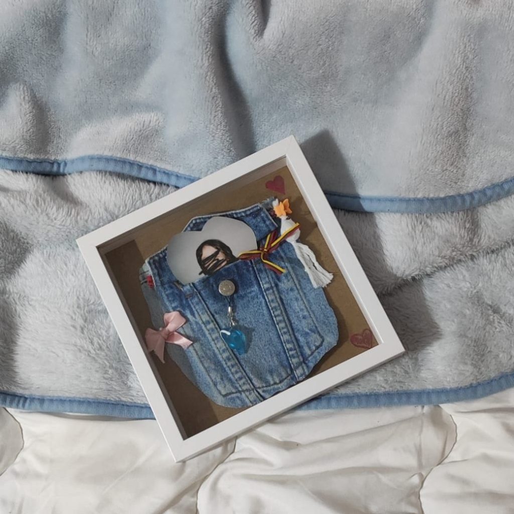 A decorative frame featuring a denim pocket with embellishments, including ribbons, a keychain, and a photo peeking out, placed on a soft blanket.
