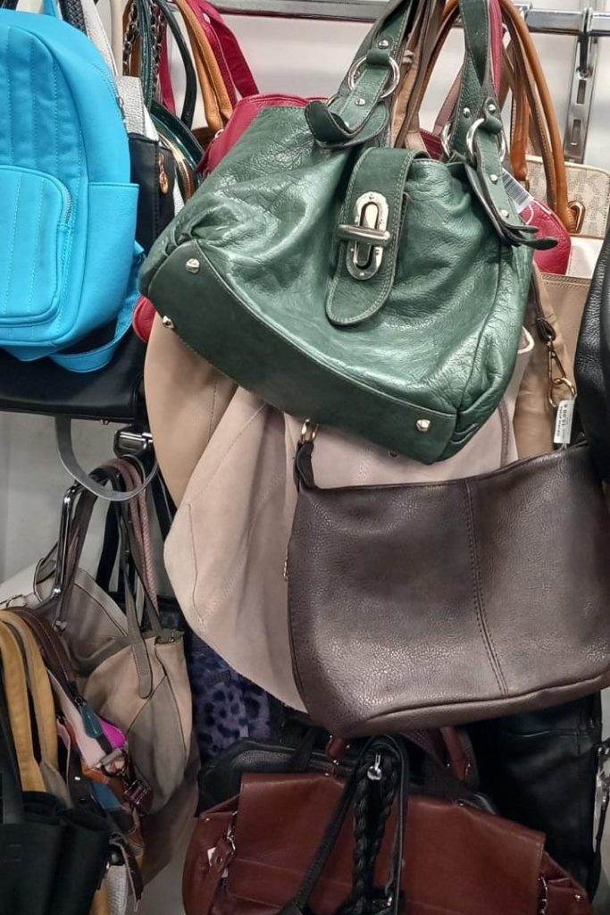 A variety of handbags and backpacks hanging on a rack, including a green leather handbag, a brown leather purse, and a blue backpack.