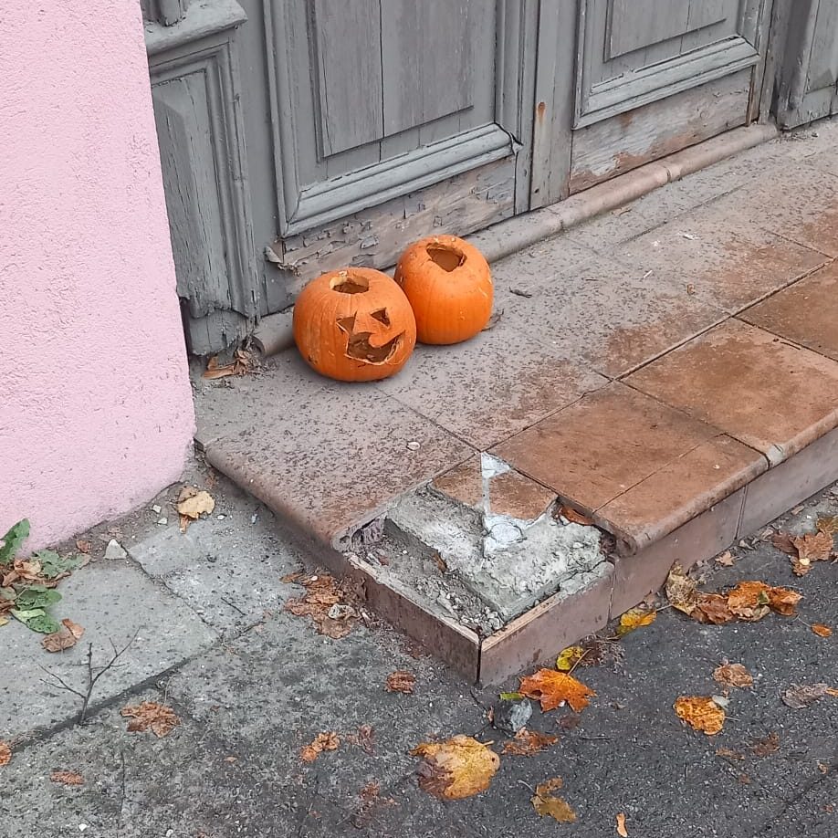 Two carved Halloween pumpkins with faces placed on a sidewalk near a weathered wooden door, surrounded by fallen leaves.