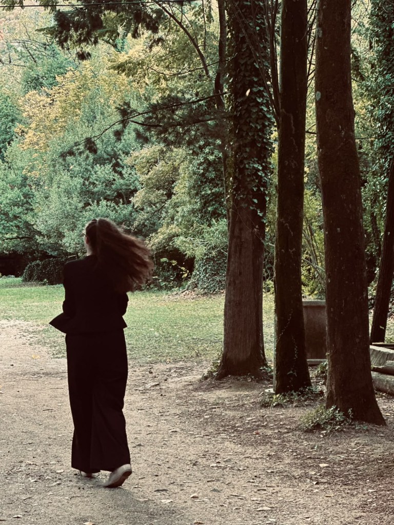 A person with long hair walks along a tree-lined path in a park, surrounded by lush greenery and tall trees.