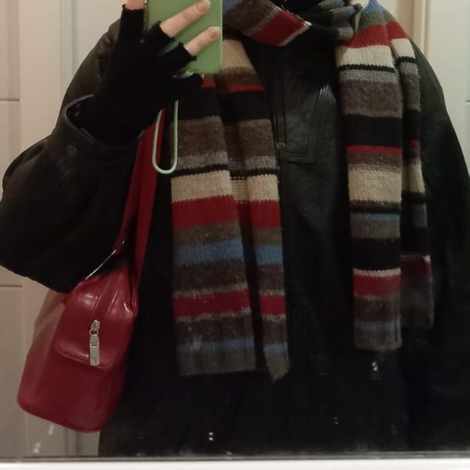 A person wearing a black leather jacket and a colorful striped scarf, holding a green phone and a red handbag.