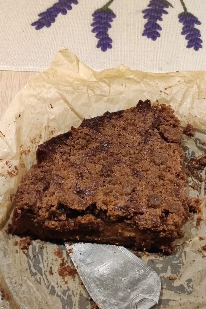 A slice of brown cake on parchment paper next to a metal spatula, with a patterned cloth in the background.