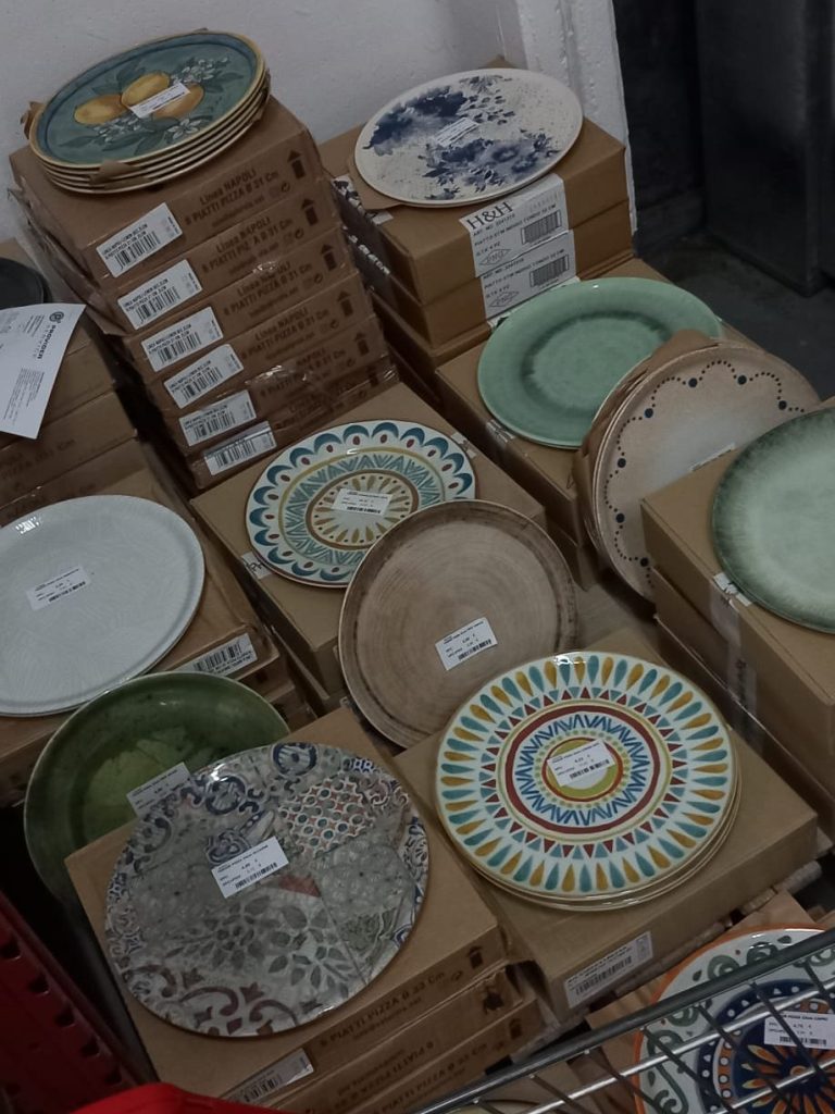 A variety of decorative plates stacked on cardboard boxes, featuring different colors and patterns. Some plates are round while others are slightly oval, showcasing intricate designs and textures.