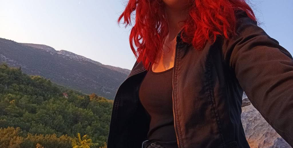 A person with red curly hair wearing a black jacket and tank top, standing outdoors with mountains and greenery in the background during sunset.