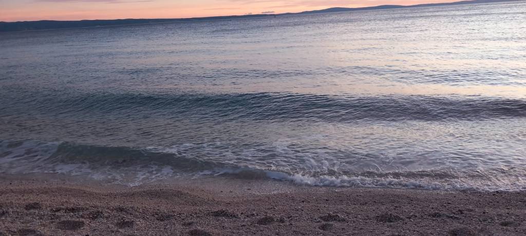 A serene beach scene showing gentle waves lapping at the shore during sunset, with a calm sea and a pastel-colored sky.
