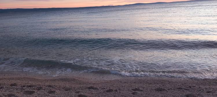 A serene beach scene showing gentle waves lapping at the shore during sunset, with a calm sea and a pastel-colored sky.