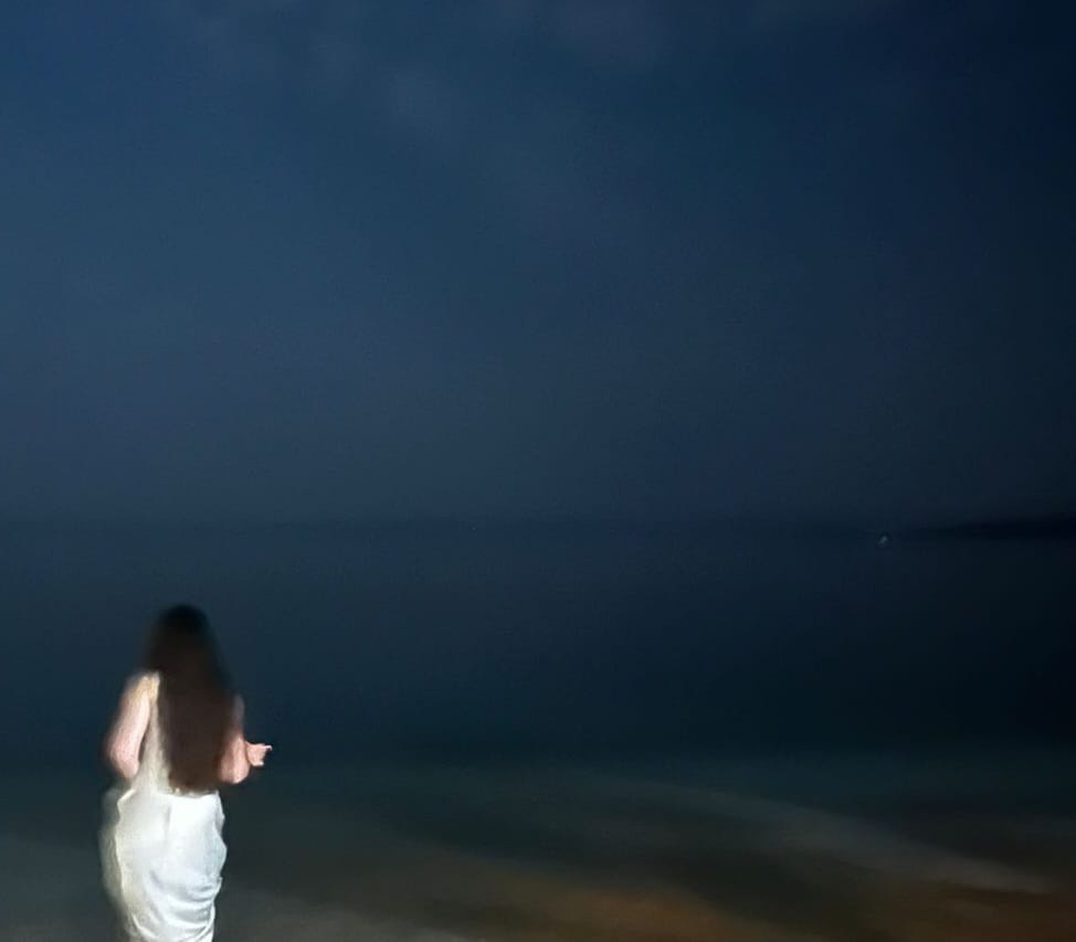 A woman with long hair, wearing a white dress, walking along the shore of a dark beach at night, with calm waters and a slightly cloudy sky.