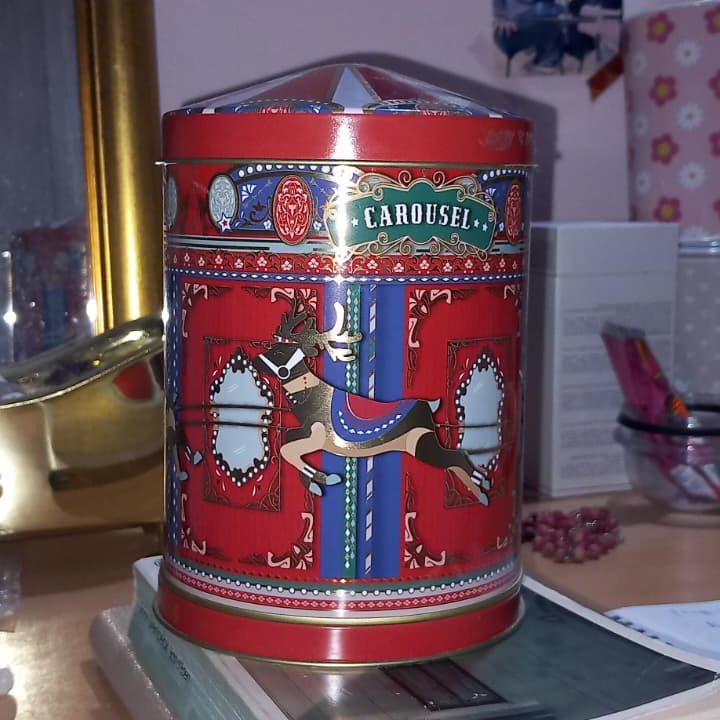 A decorative metal tin featuring a carousel theme, with a vibrant red background and illustrations of a carousel horse. The tin has a domed lid and is placed on a desk among various personal items.